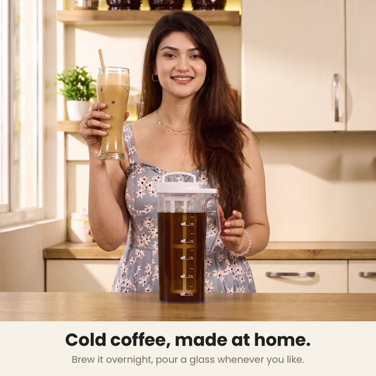 InstaCuppa cold brew coffee maker — woman holding a tall glass of Indian cafe-style cold coffee at her kitchen counter beside the jug of overnight brew