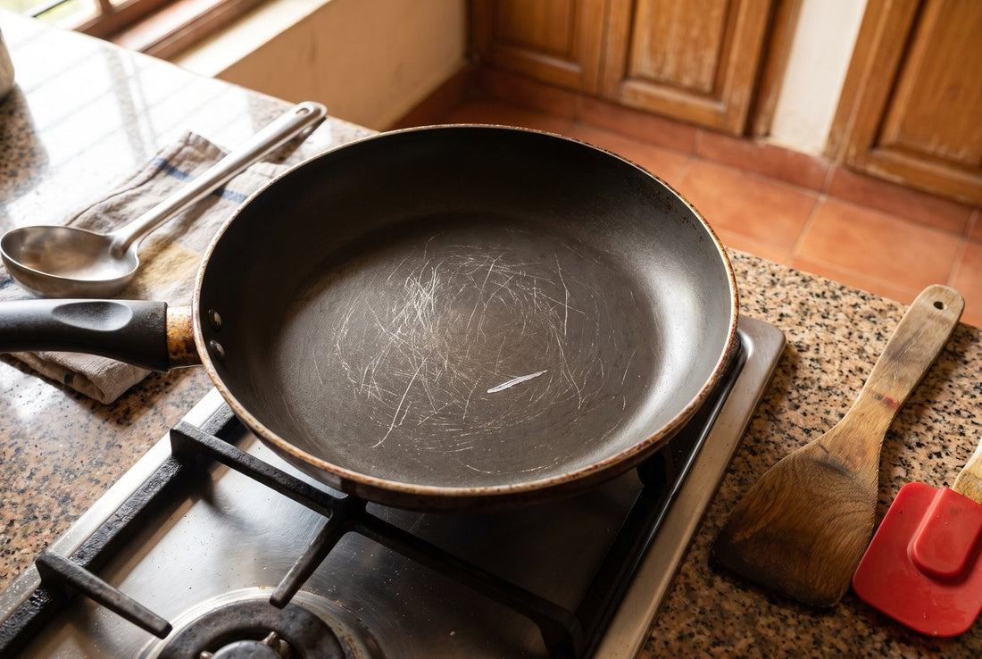 Scratched black non-stick frying pan on a gas stove, with a steel karchi on one side and a wooden spatula and red silicone spatula on the other