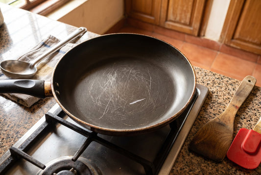 Scratched black non-stick frying pan on a gas stove, with a steel karchi on one side and a wooden spatula and red silicone spatula on the other
