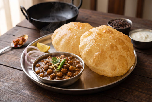 Two puffed golden bhature on a steel thali beside a katori of dark brown Punjabi chole topped with ginger strips and green chilli, with onion rings, lemon, pickle, curd and an iron kadai behind