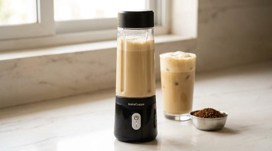 InstaCuppa portable blender with a glossy black base and clear bottle filled with frothy cold coffee on a marble Indian kitchen counter, next to a tall glass of iced cold coffee and a small steel bowl of instant coffee granules
