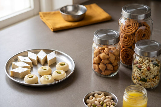 Fresh Diwali milk sweets on an open steel thali beside three sealed glass jars of dry faral holding chakli spirals, fried shankarpali diamonds and poha chivda, with cashews, pistachios and ghee alongside