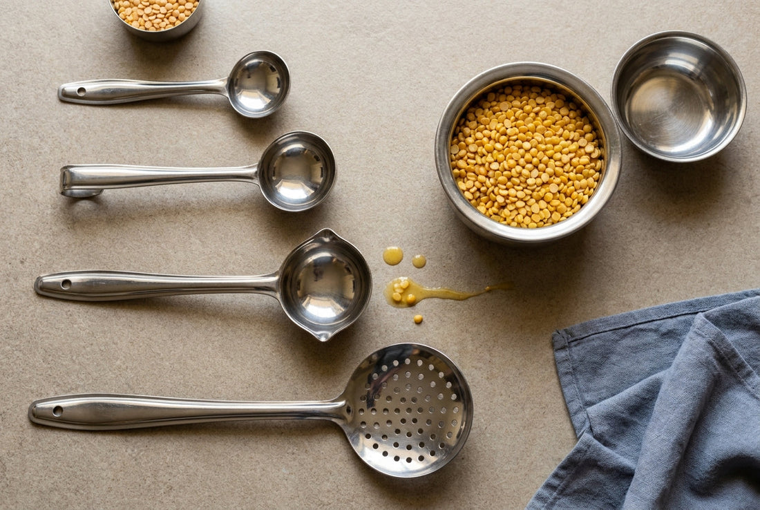 Four stainless steel ladles of increasing bowl size, one with a pouring spout and one a perforated jhara skimmer, laid beside a steel katori of yellow dal on an Indian kitchen counter