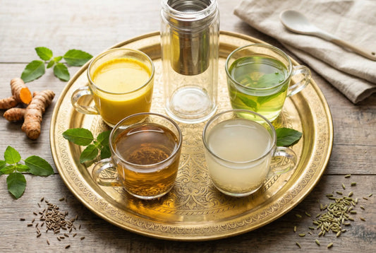 Overhead shot of 4 Ayurvedic morning drinks on a brass thali with a glass tea infuser bottle and Indian spices