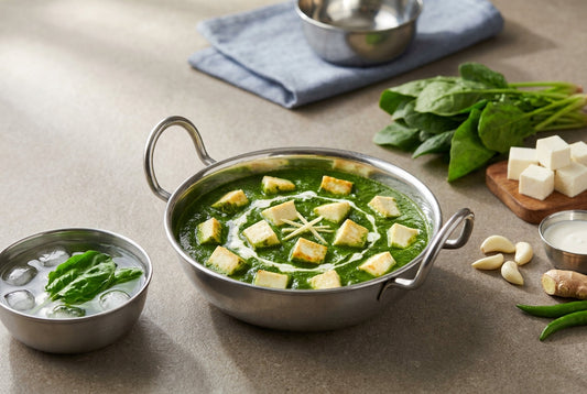Bright green palak paneer with white paneer cubes and a swirl of cream in a plain steel kadai, beside a steel bowl of ice water holding blanched spinach leaves, raw palak, garlic and ginger on an Indian kitchen counter