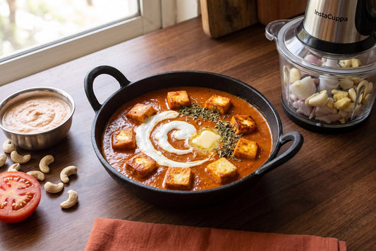 Paneer butter masala in a cast-iron kadai with a smooth unsplit swirl of cream, cashew-tomato paste in a steel katori, halved tomato and an InstaCuppa electric chopper holding onion, garlic and ginger