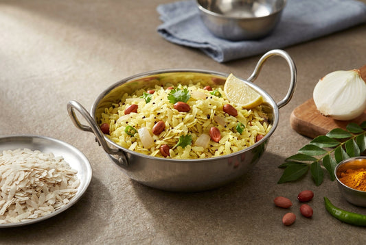 Kanda poha with separate yellow flattened rice flakes, peanuts and coriander in a plain steel kadai, beside a plate of dry uncooked poha, onion, curry leaves and turmeric on an Indian kitchen counter