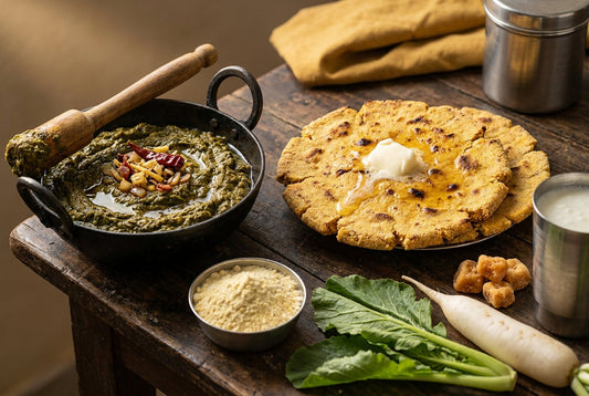 Sarson ka saag in an iron karahi with a wooden ghotni, beside two thick hand-patted makki ki rotis topped with melting white butter, with maize flour, mustard leaves, radish and jaggery on a wooden table
