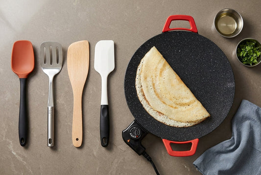 Silicone, stainless steel, wooden and nylon spatulas laid beside an InstaCuppa electric non-stick tawa with a dosa on it, on an Indian kitchen counter