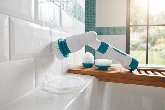 Electric spin scrubber cleaning white bathroom tiles with foam, spare brush heads on a wooden shelf