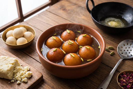 Glossy golden-brown gulab jamun in warm syrup with saffron and pistachio in a terracotta bowl, beside smooth raw dough balls, crumbled khoya, an iron kadai, a slotted spoon and rose petals