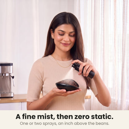 Coffee bean dosing cup held in one hand while a woman mists the beans with the RDT spray bottle, one or two sprays an inch above the beans, in a sunlit kitchen