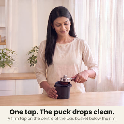 Coffee knock box in use: a woman knocks an espresso portafilter on the centre of the bar and the spent puck drops into the box in her sunlit kitchen