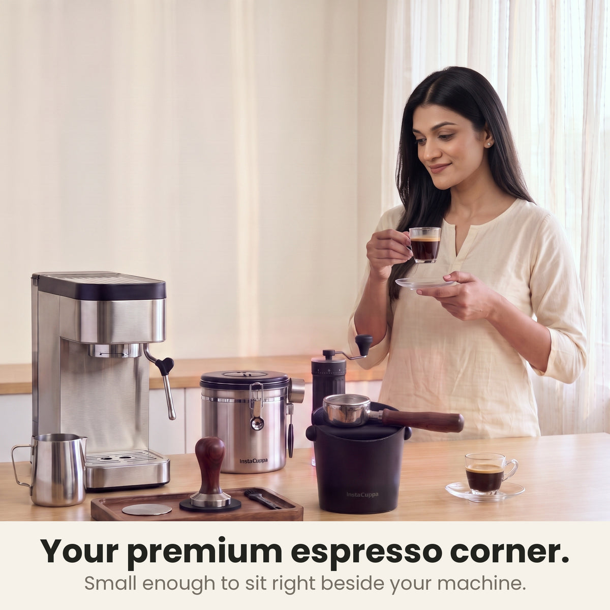 Coffee knock box in a premium home espresso corner with a slim espresso machine, tamper tray, steel coffee canister and hand grinder while a woman sips espresso