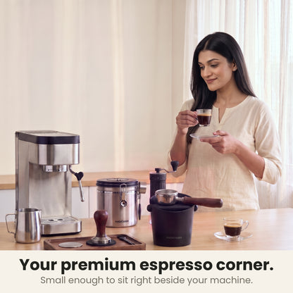 Coffee knock box in a premium home espresso corner with a slim espresso machine, tamper tray, steel coffee canister and hand grinder while a woman sips espresso