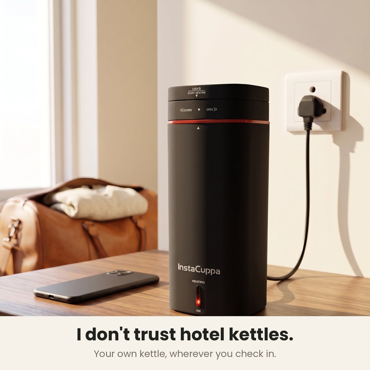 InstaCuppa Portable Electric Kettle 500 ml - Black - on a hotel desk, plugged in beside a travel bag